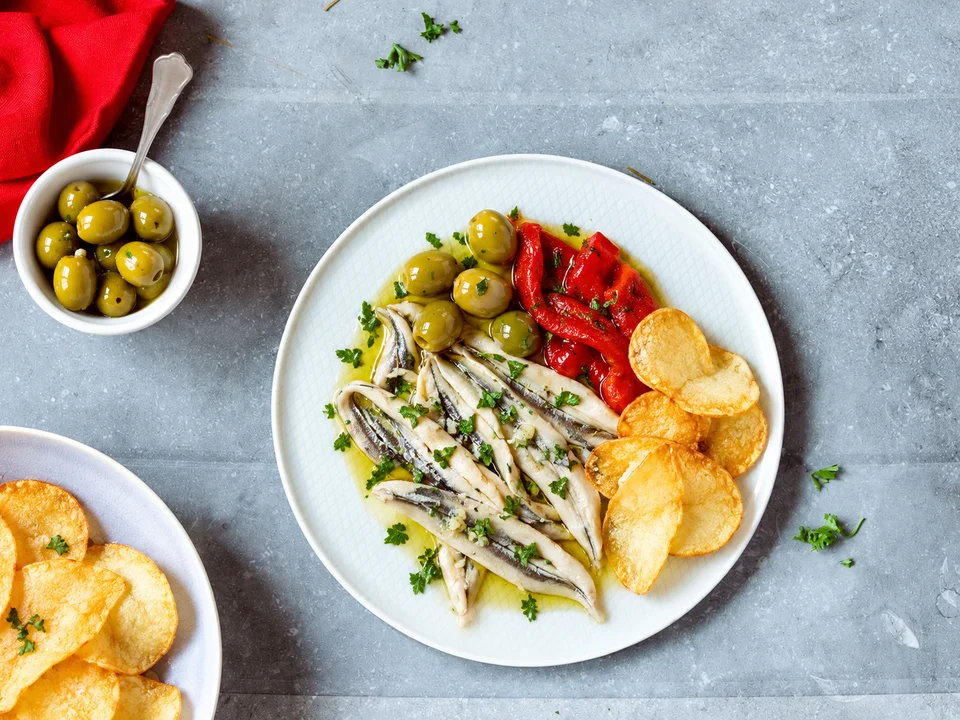 Aperitivo español de boquerones, aceitunas, pimientos y patatas fritas sobre un plato blanco.
